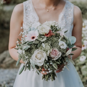 A bride in a white, lace shoulder dress, holding a bouquet of which and peach coloured flowers including roses